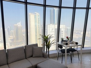 A modern hotel room with a large window offering a city view, a sofa, a small table with a vase of flowers, and a potted plant.