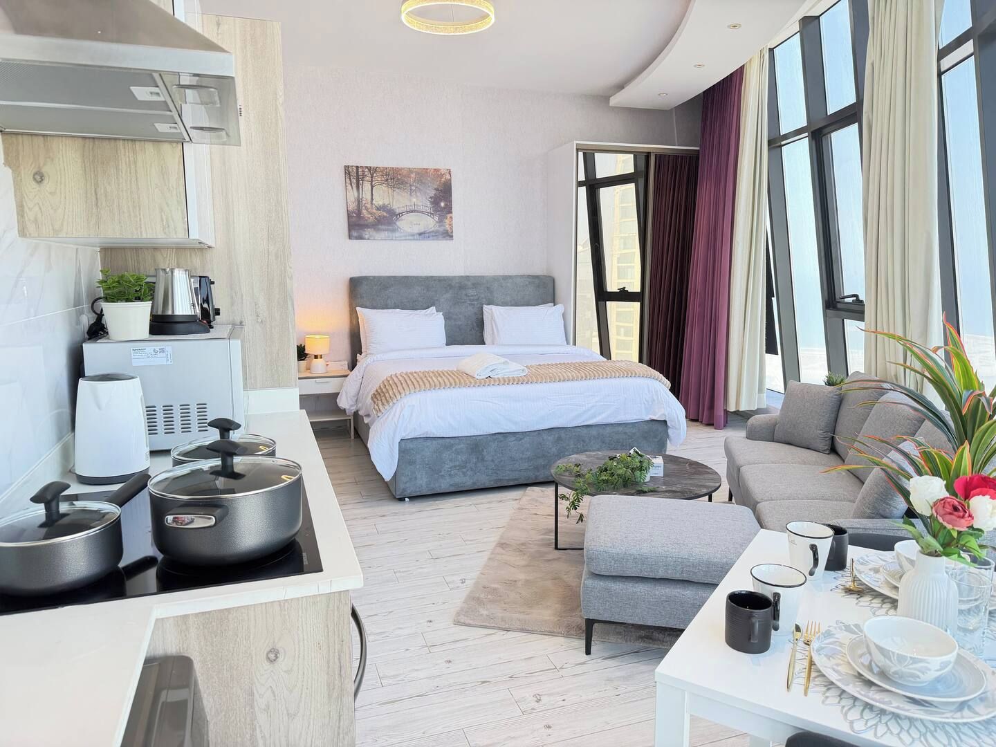 A modern hotel room with a bed, a sofa, a dining table set, and a kitchen area with appliances.