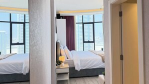 A hotel room with two beds, a desk with a lamp, and a view of tall buildings. - High-Floor Sea View Studio - Seef Area F296O (Seef)