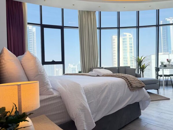 A modern hotel room with a large bed, a desk, and a view of the cityscape through floor-to-ceiling windows.