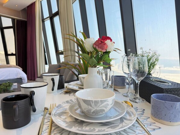 A dining table set with plates, bowls, cutlery, and glasses, with a view of the sea through large windows.