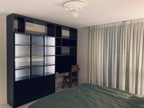 Een slaapkamer met een bed, een houten stoel, een zwart kastenrek en een raam met gordijnen.