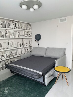 A modern bedroom with a bed, a wall-mounted shelf with decorative items, a round table with a yellow top, and a grey sofa. -  Luxury Designer-Furnished 2BR with 2 Large Terraces in Bay Harbor (Bay Harbor Islands)