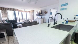 A modern living room with a sofa, a dining area, and a kitchen with a large island and a sink. - Saida Royale, Beachfront 10th Floor, 2BR, 3 Pools (South Padre Island)