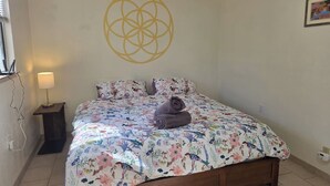 Ein Schlafzimmer mit floralem Bettbezug, einem hölzernen Bett, einer Wandlampe und einer geometrischen Wanddekoration.