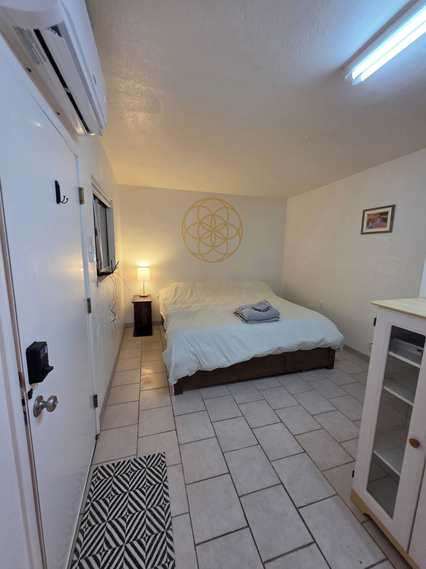 A bedroom with a bed, bedside lamp, air conditioning unit, and a geometric wall decoration.