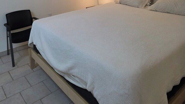 Ein Schlafzimmer mit einem Bett, einem Sessel, einer Lampe und einem Fenster mit Jalousien.