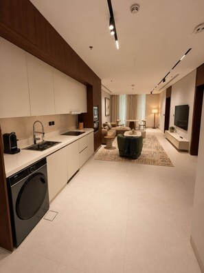 A modern kitchen with a built-in washer and dryer, a dining area with a sofa and a coffee table, and a living room with a TV and a lamp. - LIV by Yamama - Park 91 (Riyadh)