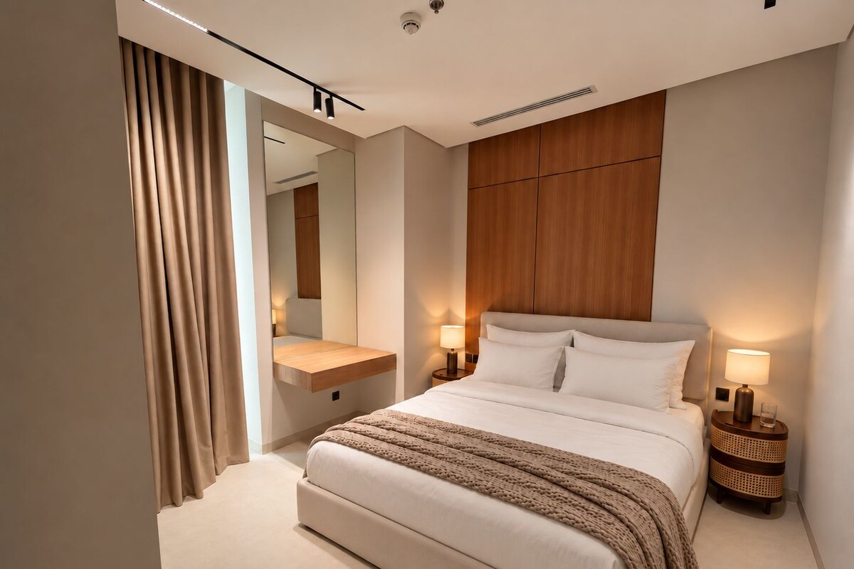 A modern hotel room with a large bed, bedside lamps, a wooden headboard, a mirror, and a bedside table.
