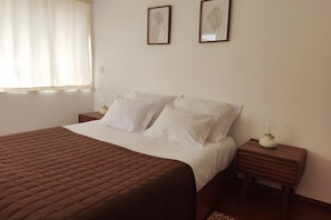 Cama bem arrumada com colcha marrom, lençóis brancos e duas mesas de cabeceira com abajures.