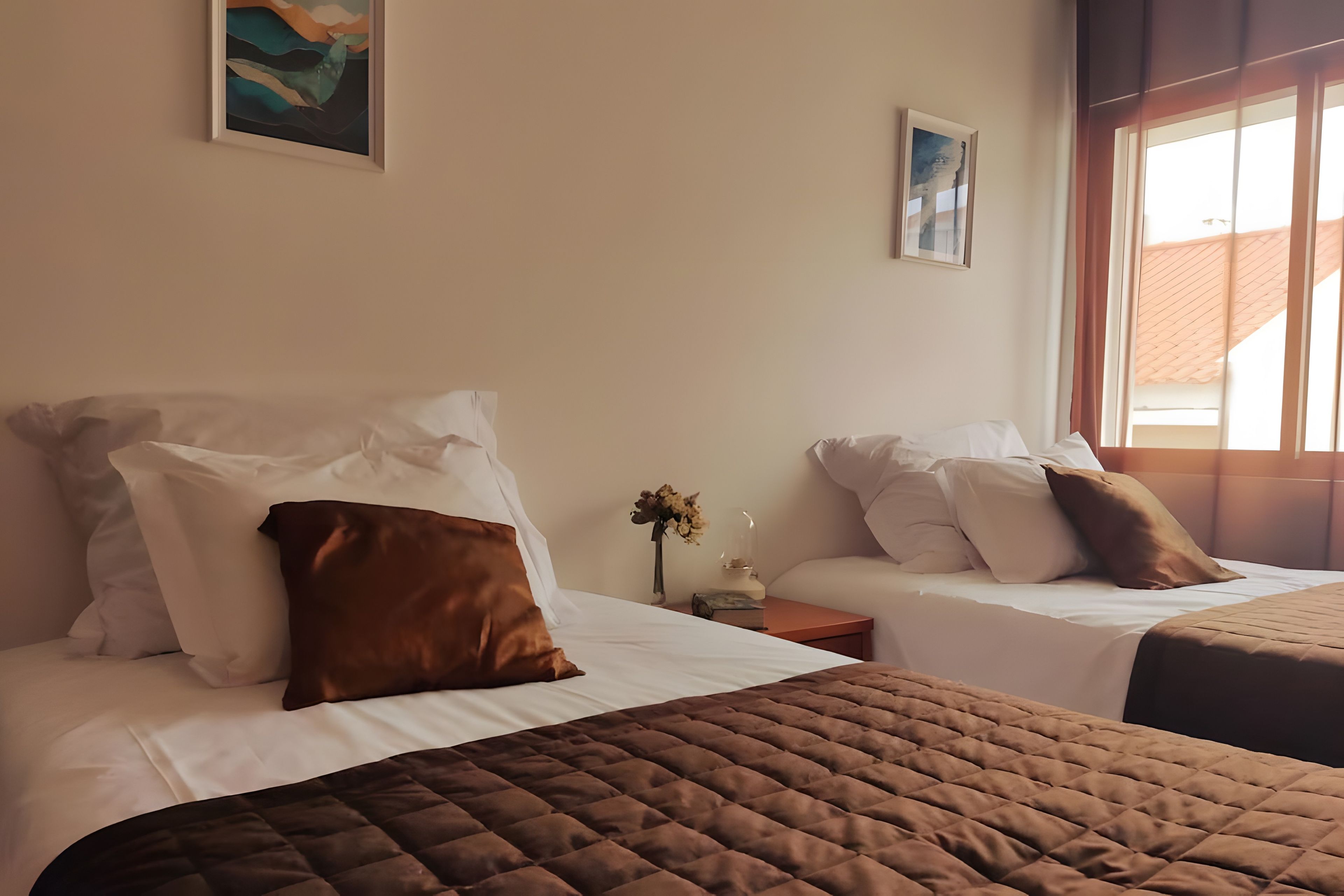 Habitación de hotel con dos camas, cada una con ropa de cama blanca y almohadas marrones. Se ve una ventana al lado derecho, y hay obras de arte en las paredes.