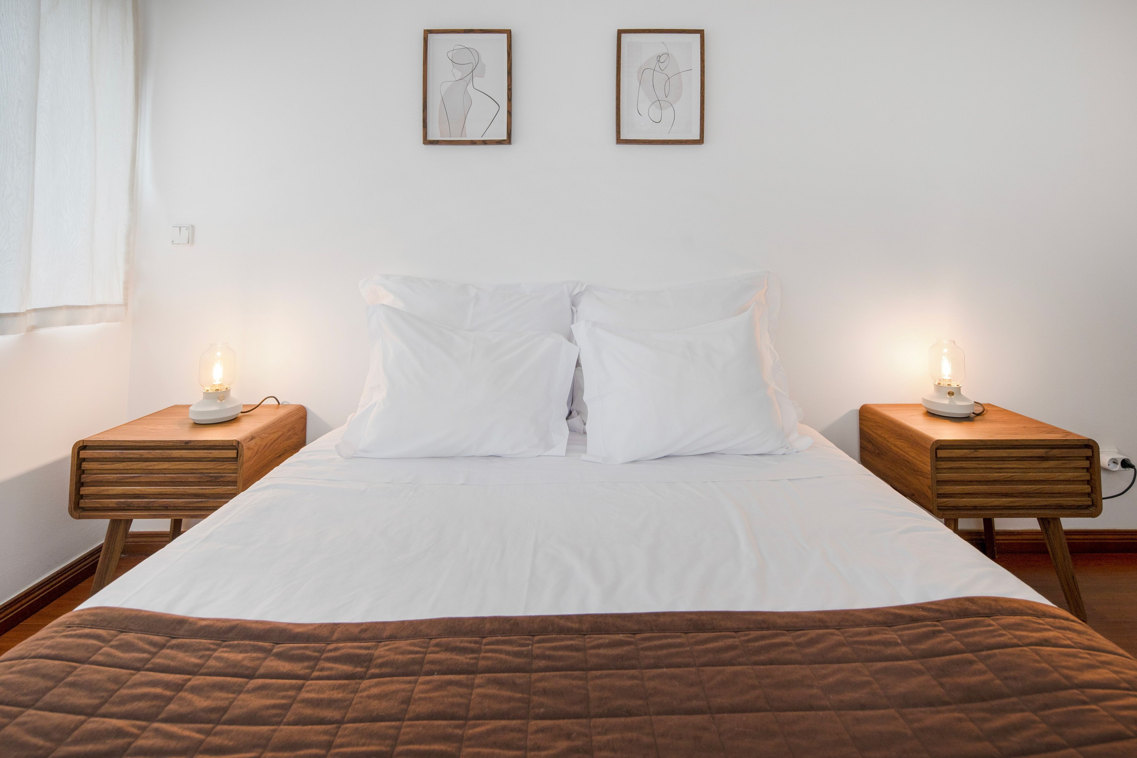 A bedroom with a bed, two bedside tables, two wall-mounted picture frames, and a window with curtains.