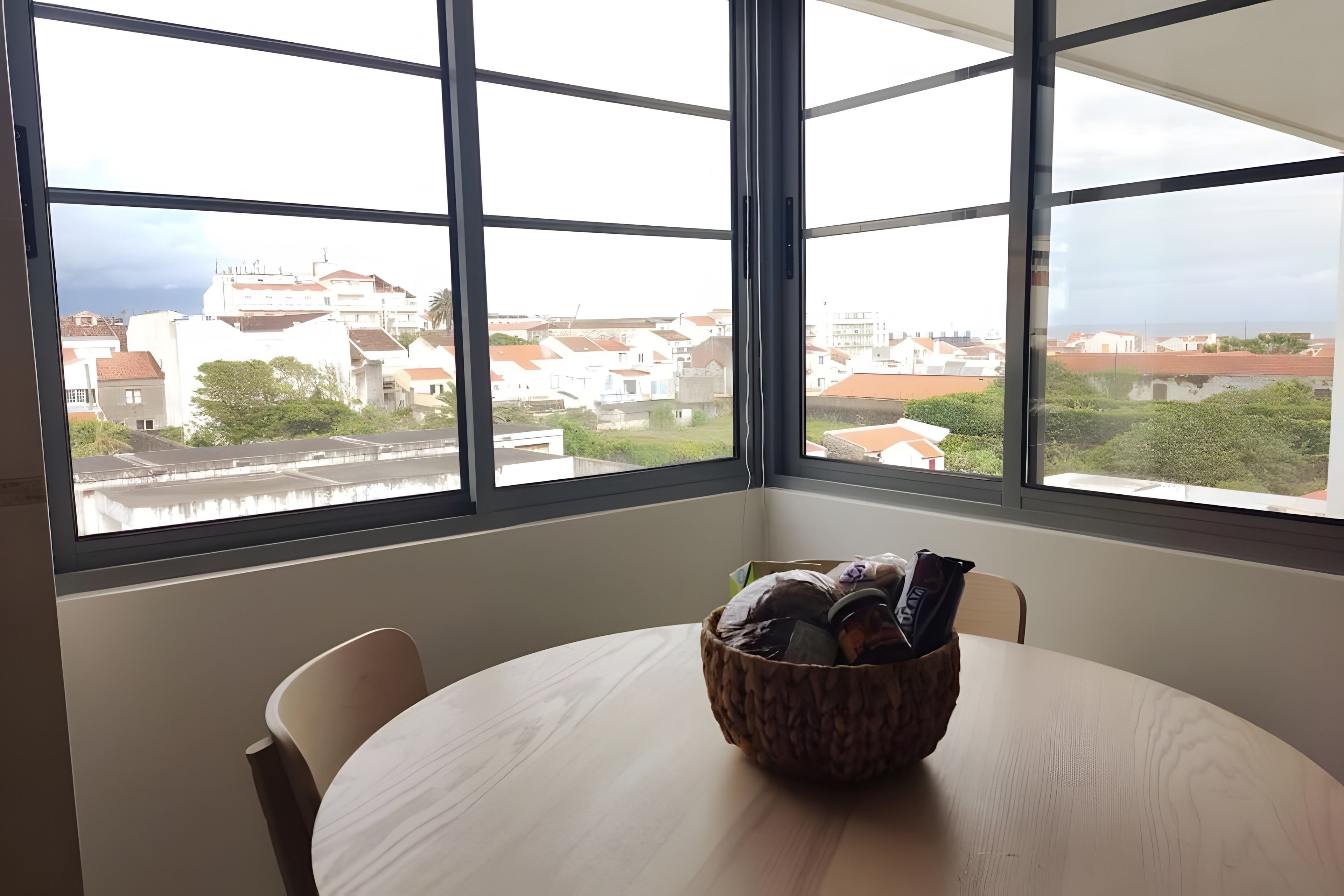 Una habitación con una mesa redonda y sillas, una ventana que ofrece vistas a edificios, y una cesta sobre la mesa.