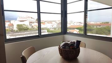 Una habitación con una mesa redonda y sillas, una ventana que ofrece vistas a edificios, y una cesta sobre la mesa.