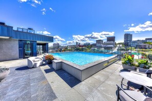 Ein Pool auf dem Dach mit Blick auf die Stadt, ein modernes Gebäude mit blauer Fassade und Außenbereich mit Sitzmöglichkeiten.