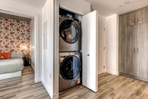 A laundry room with two stacked front-loading washing machines, a wooden wardrobe, and a bed with a patterned wall in the adjacent room. - El Emilio · Parking · Piscine et spa · 2 chambres (Quebec City)