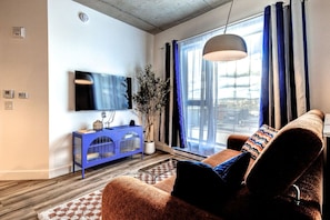 A modern living room with a blue console, a large window with curtains, a sofa with patterned cushions, and a hanging light fixture. - El Emilio · Parking · Piscine et spa · 2 chambres (Quebec City)