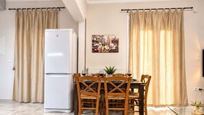 Un comedor con un refrigerador blanco, sillas de madera, una mesa de vidrio y cortinas color beige.