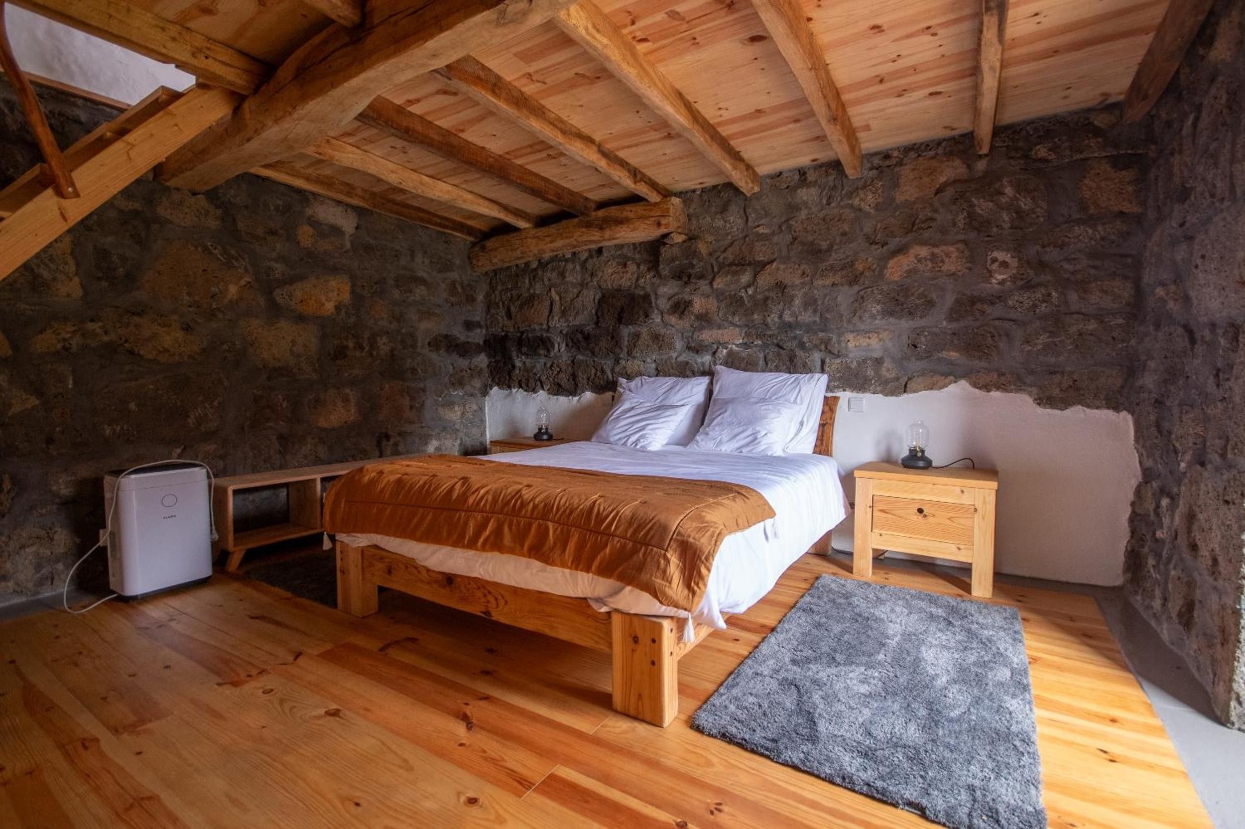 Une chambre avec un mur en pierre, des poutres en bois au plafond, un lit, des tables de chevet et un radiateur.