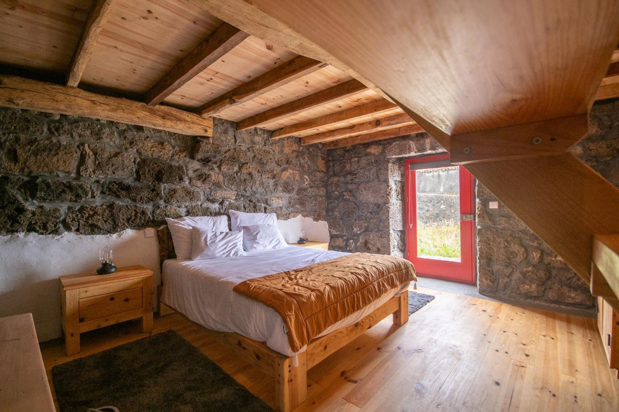 Et soveværelse med stenvæg, trælofter, en seng med hvide sengetæpper, et natbord og en rødlakeret dør.