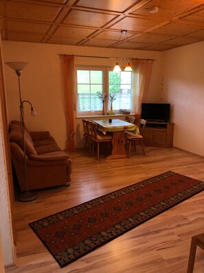 Ein gemütliches Wohnzimmer mit einer Couch, einem Esstisch mit Stühlen, einem Fernseher und einem großen Fenster mit Vorhängen.