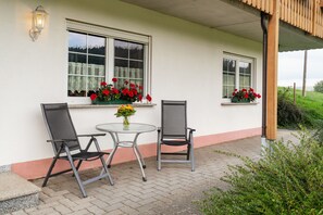 Ett takförsett utomhusområde med sittplatser, bestående av två stolar och ett bord, två fönster med blomlådor och en utsikt över grönskan i bakgrunden.