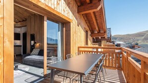 A wooden deck with a table and chairs, a sofa, and a kitchen area with a window. - Altamira – Views| Terrace| Hammam| Alpe d’Huez (Huez)