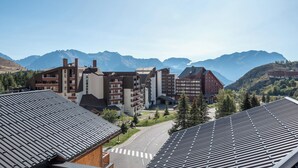 A modern hotel with multiple floors, a parking area, and a view of mountains in the background. - Altamira – Views| Terrace| Hammam| Alpe d’Huez (Huez)