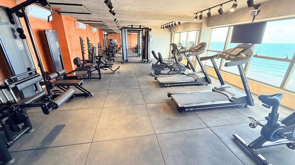 Un gimnasio con diverso equipo de ejercicio, incluyendo cintas de correr y máquinas de pesas, con vista al océano a través de grandes ventanales.
