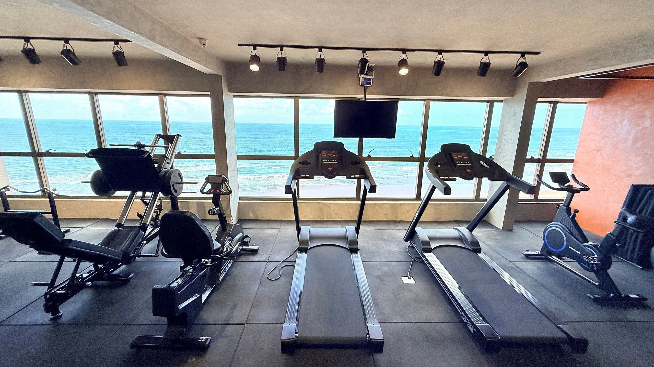 Academia com esteiras, remo e bicicleta ergométrica, com vista para um corpo de água.