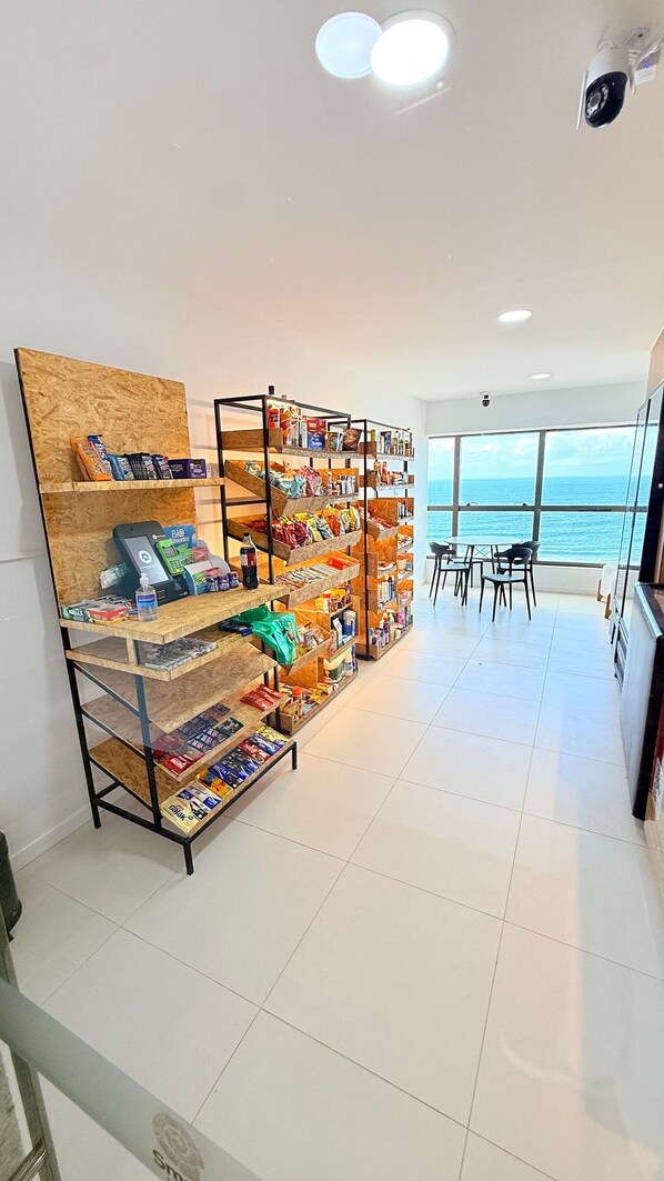 A room with a large window overlooking the sea, a bookshelf filled with various items, and a dining area with chairs.