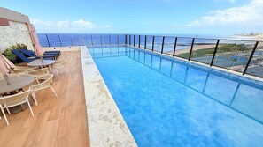 A rooftop infinity pool with a view of the ocean, lounge chairs, and a seating area with tables.