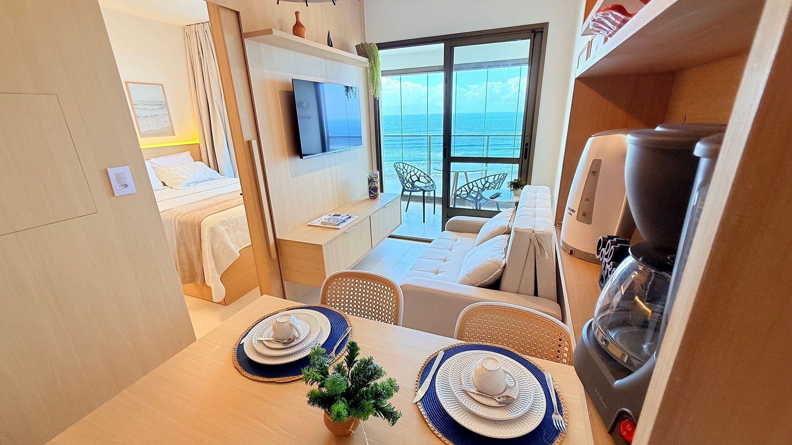 Quarto de hotel compacto com cama, área de refeições com mesa posta para dois, máquina de café e uma grande janela com vista para o mar.