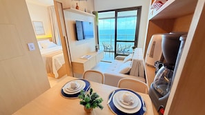 Habitación de hotel compacta con una cama, zona de comedor con mesa para dos personas, máquina de café y un gran ventanal con vistas al mar.