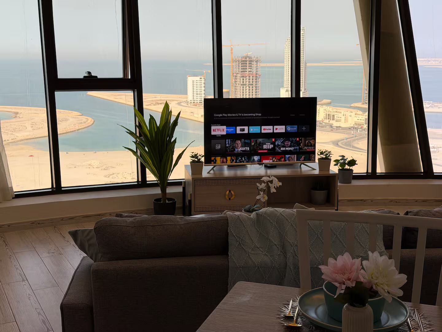 A living room with a large TV, a sofa, a dining table with a vase of flowers, and a view of a coastal cityscape.
