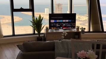 A living room with a large TV, a sofa, a dining table with a vase of flowers, and a view of a coastal cityscape.