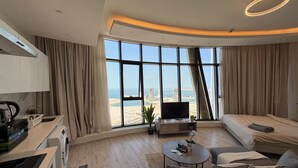 A modern hotel room with a large window, a flat-screen TV, a sofa, a coffee table, and a kitchen area. - Lovely Sea View Studio in Downtown F285O (Seef)