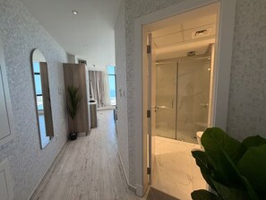 En hotelgang med glasdør til brusebad, et spejl og en potteplante.