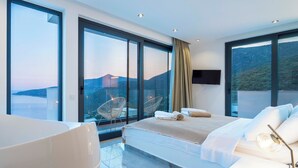 A modern hotel room with a large bed, a seating area with chairs, a television, and a balcony with a view of the sea and hills. - Vrbo Property (Kalkan)