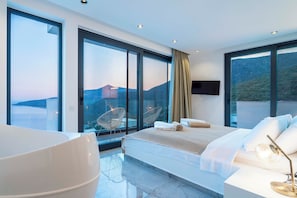 Quarto de hotel moderno com uma cama grande, área de estar com cadeiras, televisão e varanda vista para o mar e colinas.