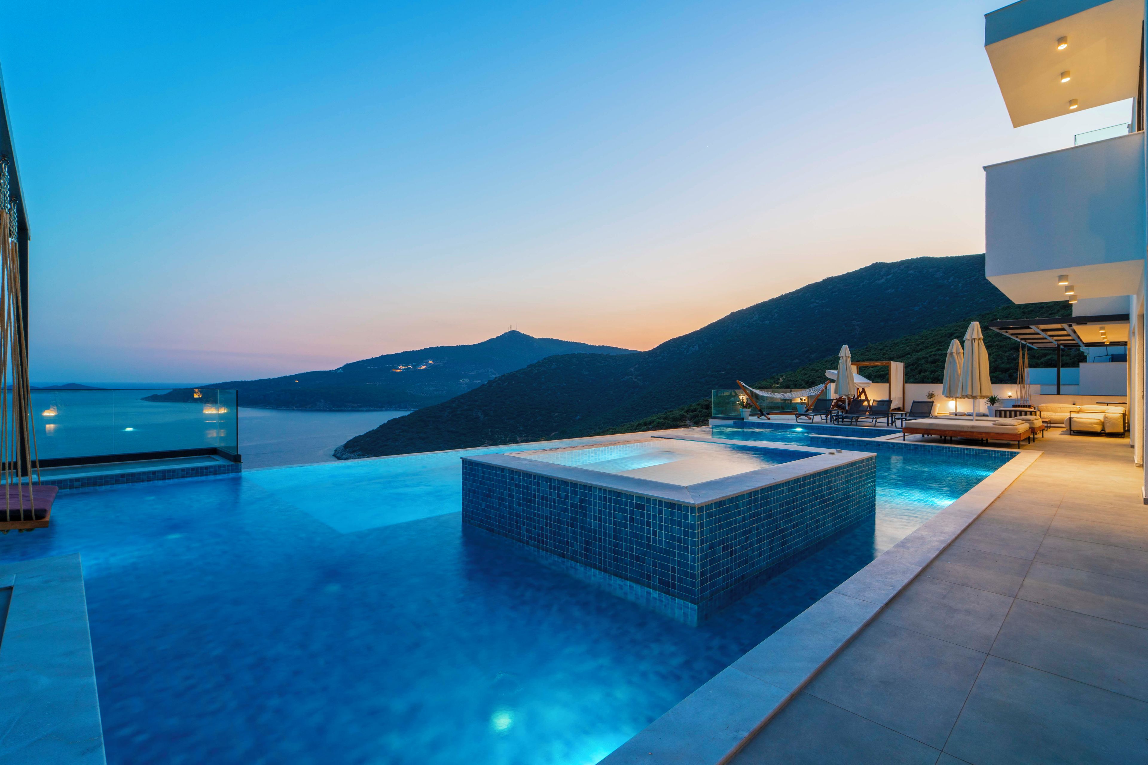 A modern infinity pool with a view of the sea and mountains at dusk.