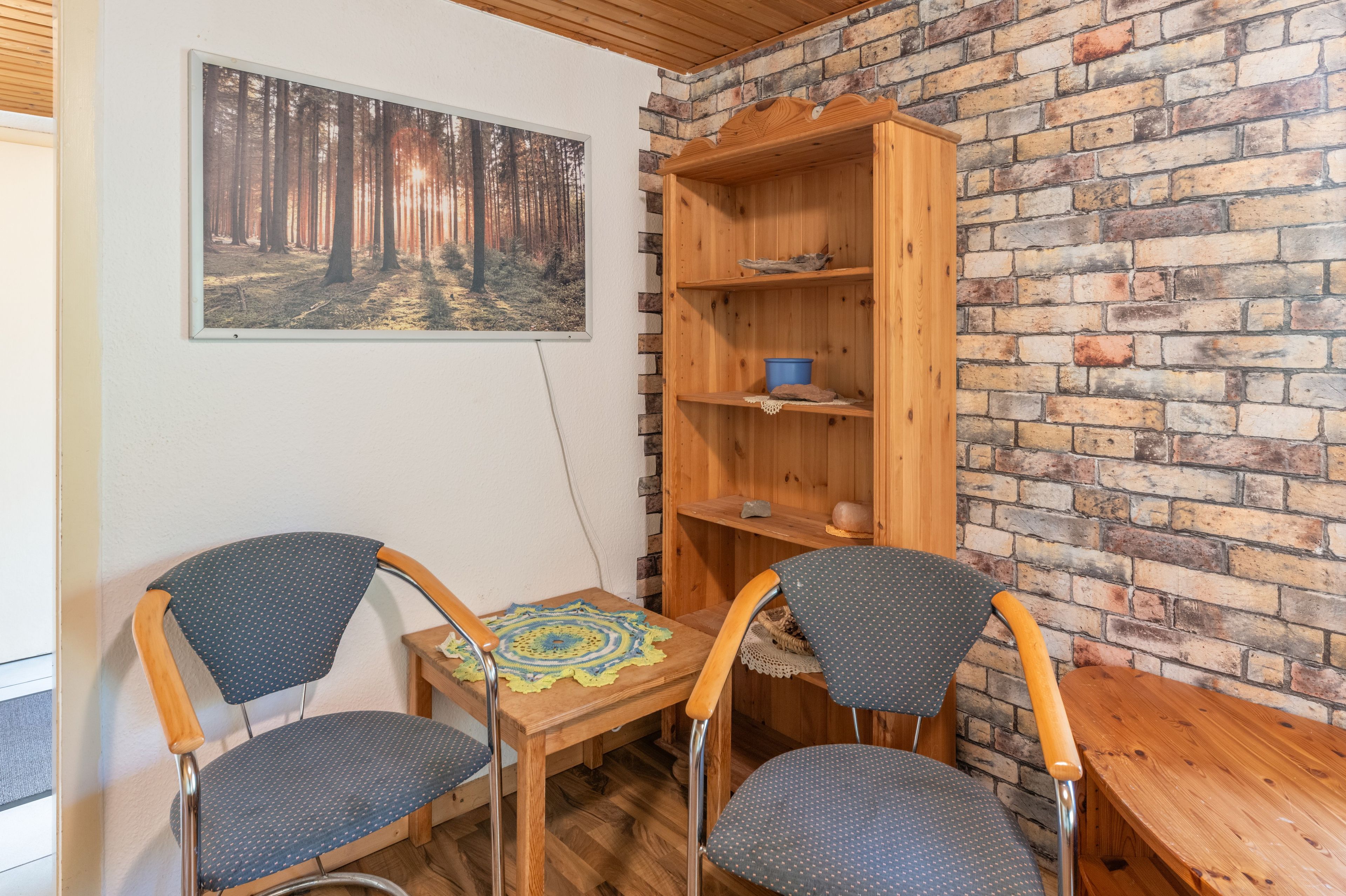 Ein Zimmer mit Backsteinwand, zwei Stühle, einem kleinen Tisch mit Häkeldecke, einem Holzregal mit blauer Schüssel und einem gerahmten Foto von einem Wald.
