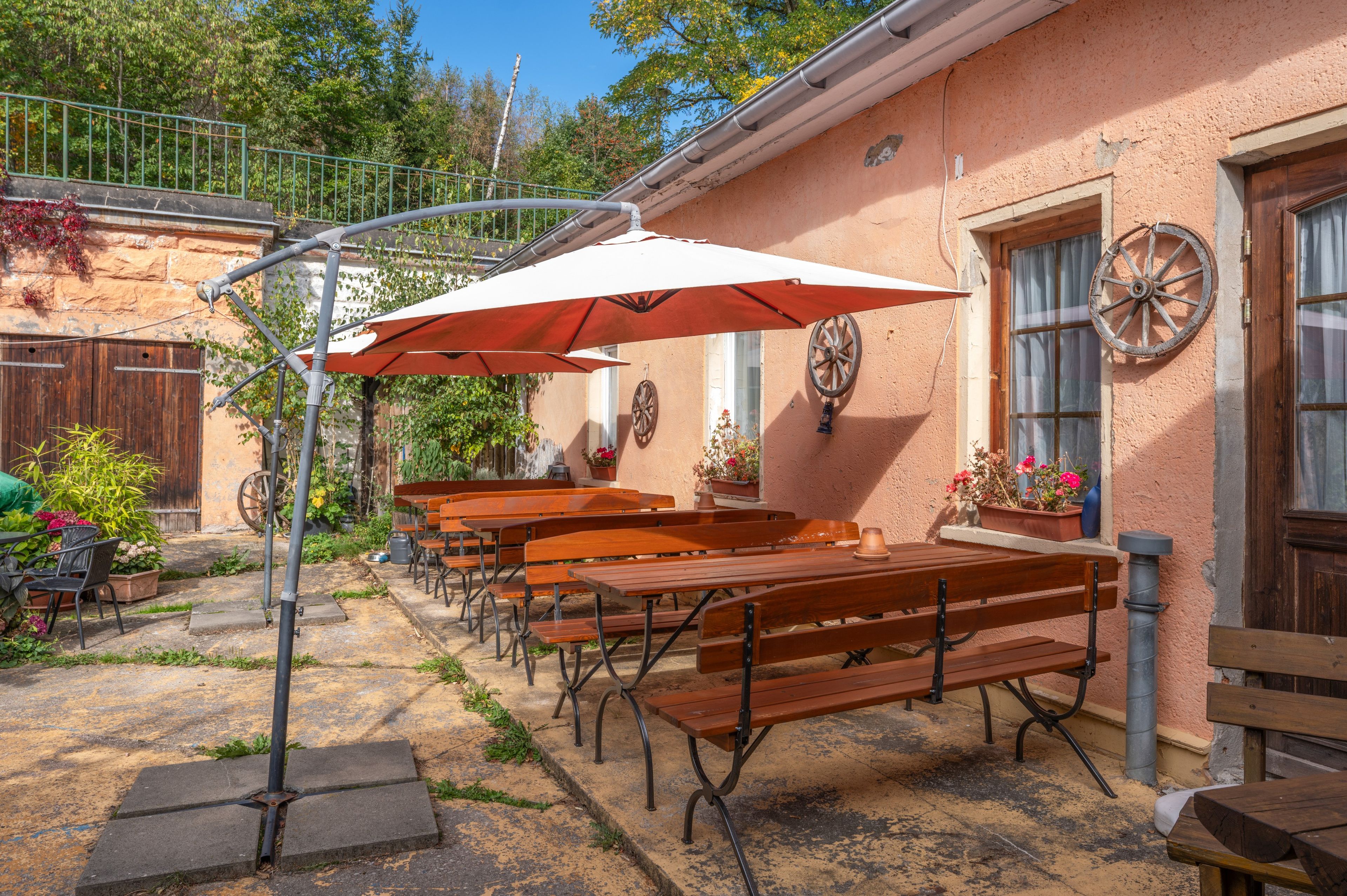 Un espace extérieur aménagé avec des bancs et des tables en bois, des parasols et un bâtiment rustique orné d’une roue sur le mur.