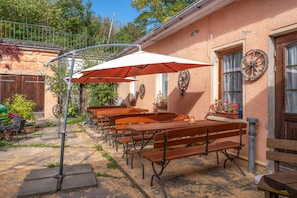 An outdoor seating area with wooden benches and tables, umbrellas, and a rustic building with a wheel on the wall.