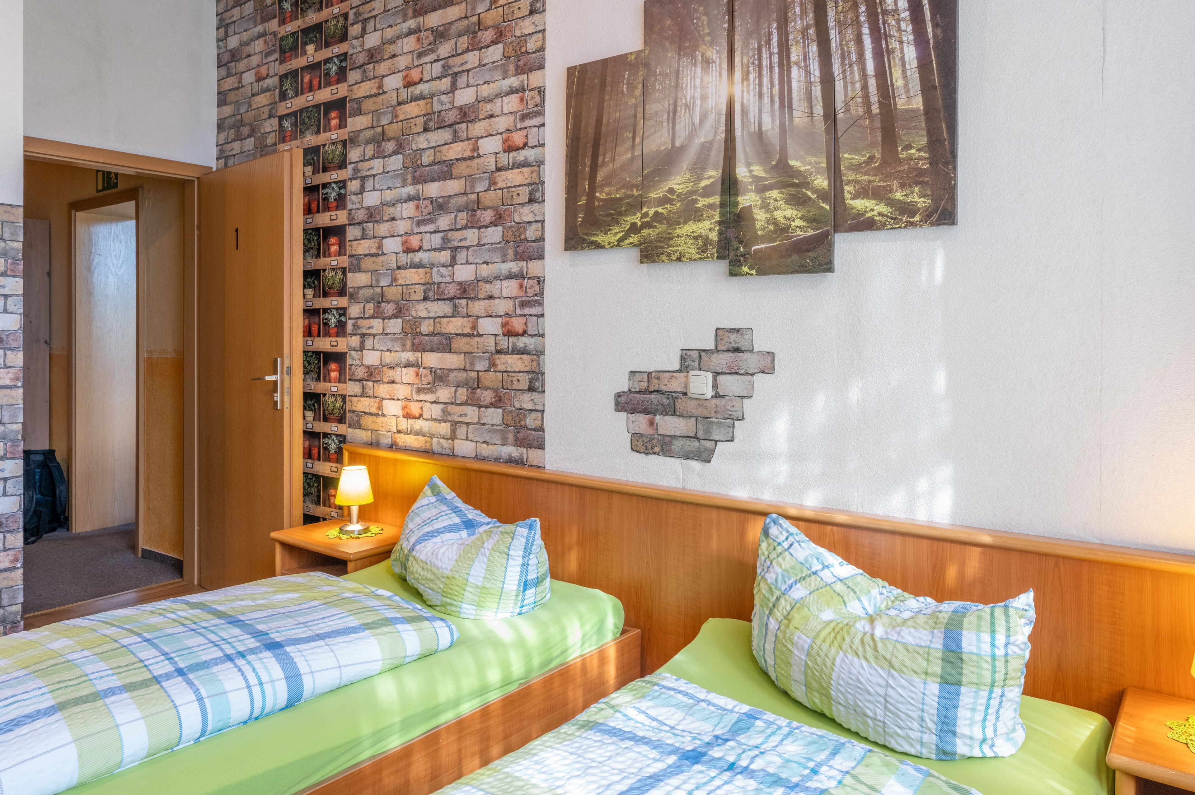 Een hotelkamer met een bakstenen muur, een bed met groene beddengoed, een nachtkastje met een lamp en een wandplank met planten.