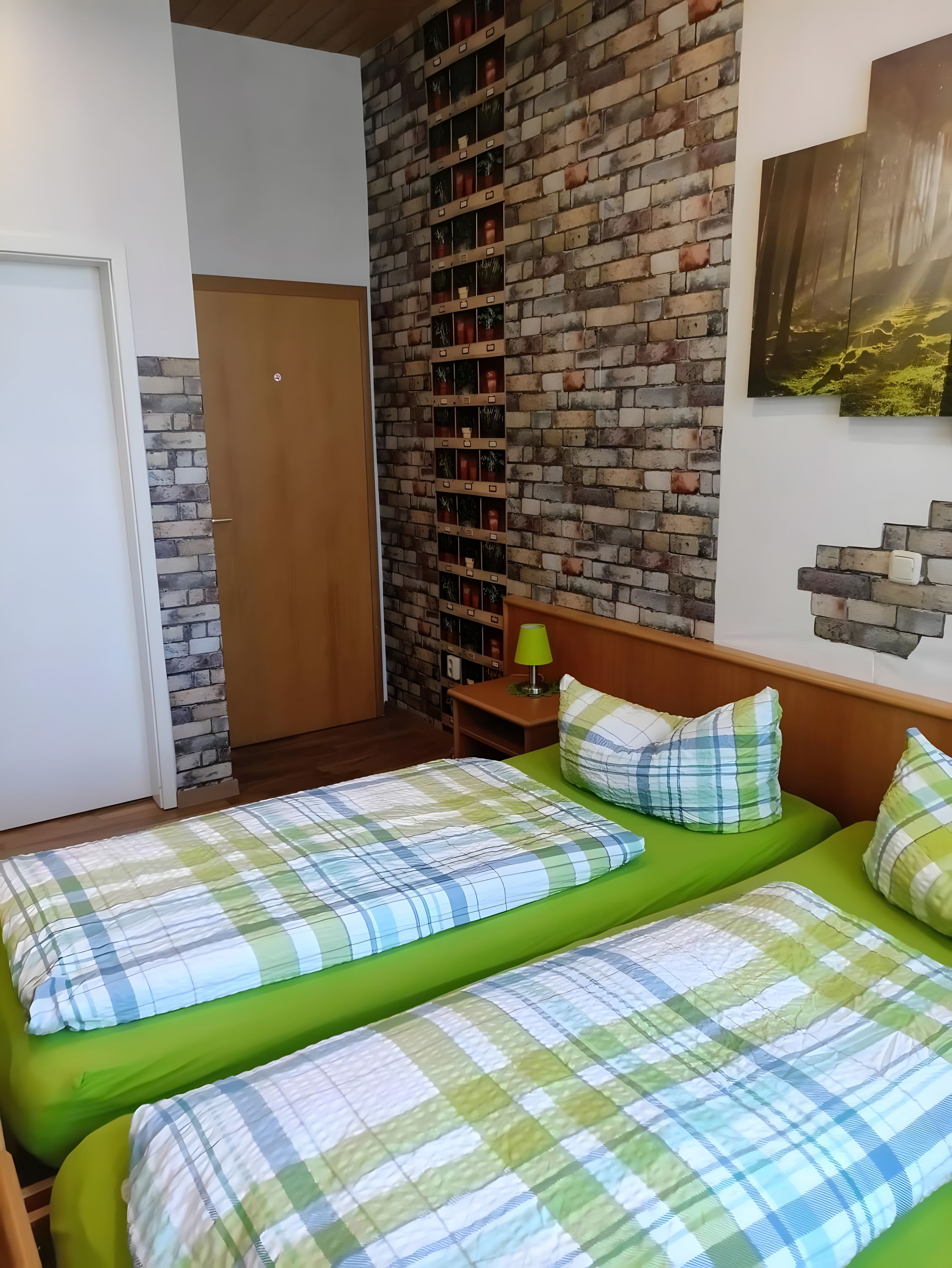 A room with two beds, a brick wall, a wooden door, and a small table with a lamp.