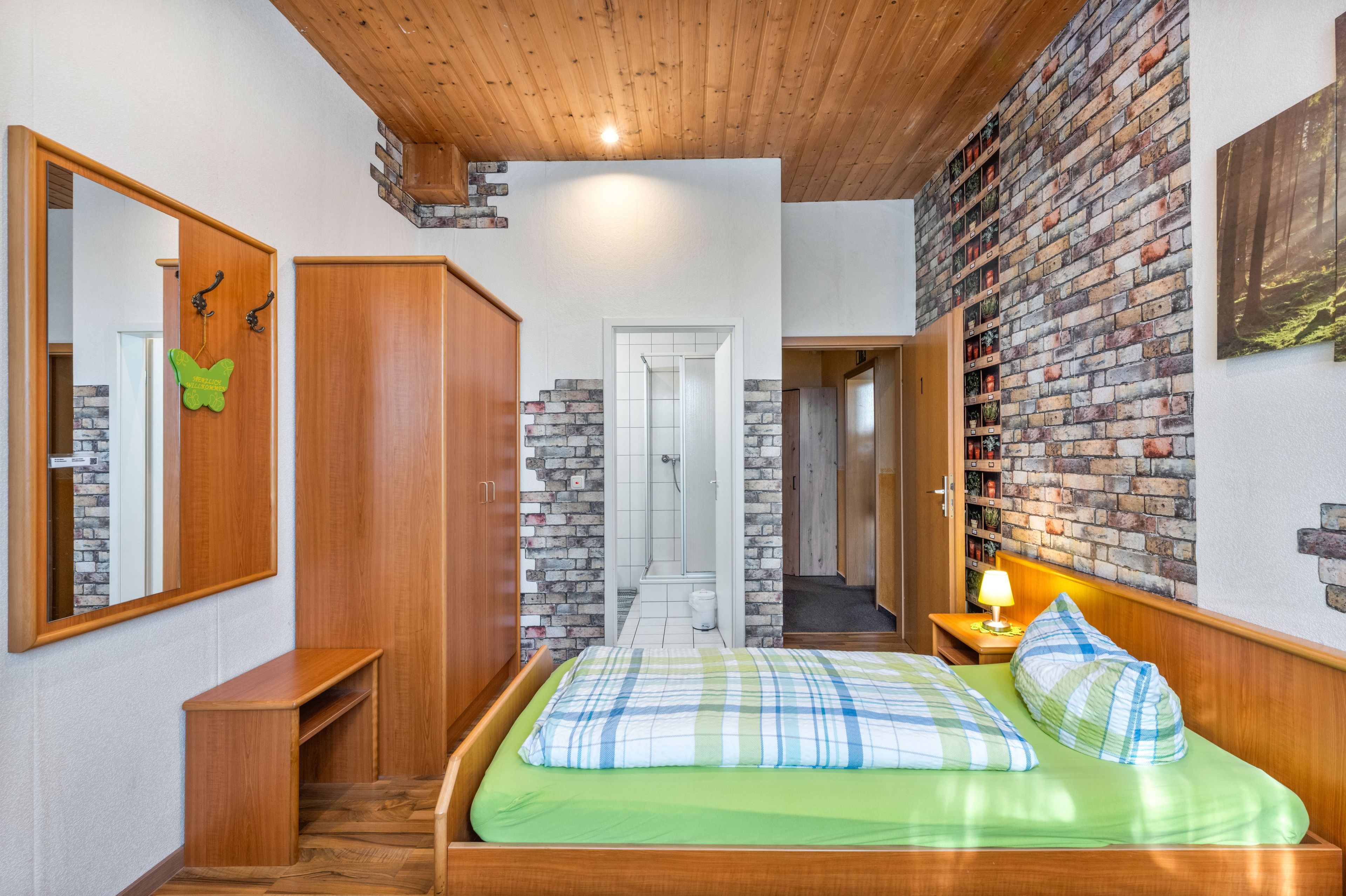 Quarto com teto de madeira, parede de tijolos, cama com roupa de cama verde, mesa de cabeceira com abajur e guarda-roupa.
