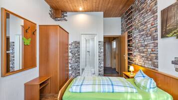 Quarto com teto de madeira, parede de tijolos, cama com roupa de cama verde, mesa de cabeceira com abajur e guarda-roupa.