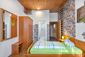 A bedroom with a wooden ceiling, a brick wall, a bed with green bedding, a nightstand with a lamp, and a wardrobe.