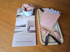 Un folleto con el título 'SUPER HOST BRASIL', que muestra un paisaje urbano y una cama, una taza azul con paquetes de chocolate y una bandeja de madera con un cuaderno y chocolate.